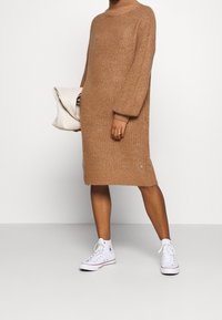 Abito in maglia oversize di colore marrone chiaro caldo. Presenta un colletto alto e maniche a palloncino. Abbinato a sneakers bianche alte.