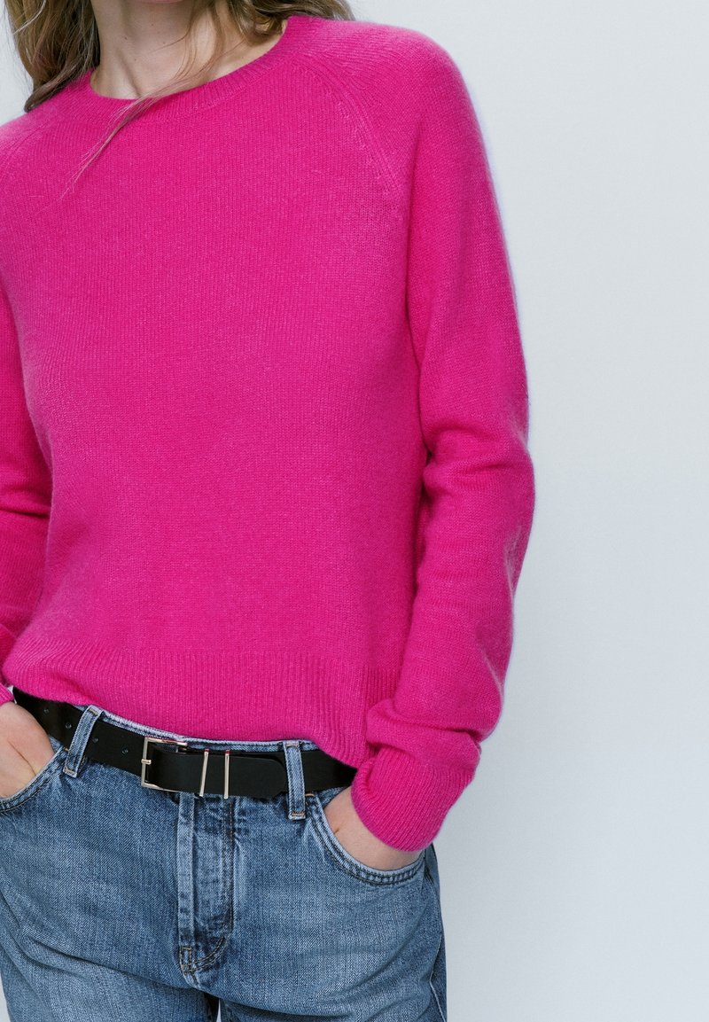 Pull fuchsia à col rond et poignets côtelés, associé à un jean bleu délavé et une ceinture noire avec bouclerie en argent.
