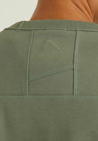 Chemise vert olive avec un col côtelé, des coutures texturées et un petit détail triangulaire cousu à l'arrière. Tissu lisse.