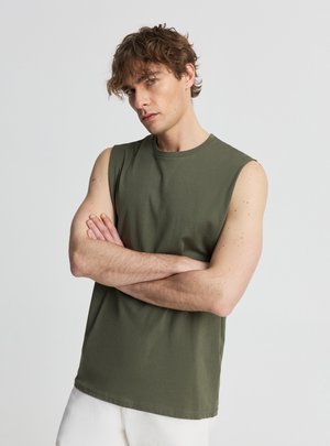 Uomo con capelli ricci castani che indossa una canotta verde oliva e pantaloni bianchi, con le braccia incrociate, che guarda leggermente di lato.