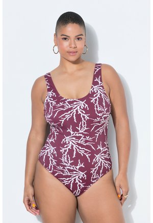 Nuori nainen, jolla on lyhyet hiukset, pukeutuneena violettiin yhtenäiseen uima-asuun, jossa on valkoisia korallinkaltaisia kuvioita, ja kultaiset korvarenkaat.