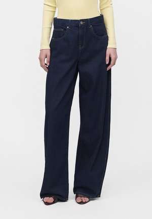 Person iført mørkeblå jeans med vide ben og en lysegul langærmet top, stående med hænder afslappet langs siden.