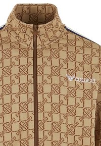 Beige zip-up jacket with a textured pattern displaying interlocking letters in brown. Features a stand collar and contrasting navy stripes.