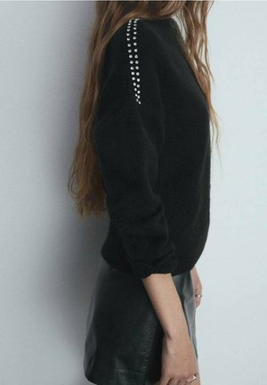 Maglione nero lavorato a maglia con maniche a sbuffo e strass decorativi sulla spalla, abbinato a una gonna in pelle, mostrato di profilo.