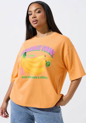 Młoda kobieta o długich ciemnych włosach, ubrana w luźny pomarańczowy T-shirt z grafiką bananów i napisem "Naturalnie słodka" oraz niebieskie dżinsy.