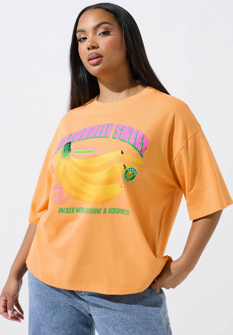 Młoda kobieta o długich ciemnych włosach, ubrana w luźny pomarańczowy T-shirt z grafiką bananów i napisem "Naturalnie słodka" oraz niebieskie dżinsy.