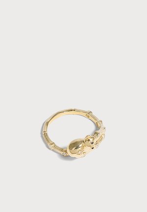 Goldener Ring mit Babyfigur und kleinen eingelassenen Steinen auf einem strukturierten Band, gezeigt vor hellem grauem Hintergrund.