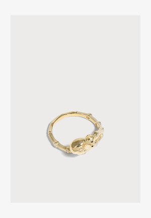 Bague en or avec une figurine de bébé et de petites pierres incrustées sur une bande texturée, présentée sur un fond gris clair.