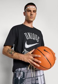 Férfi fekete "Unique" Nike pólóban és fejpántban narancssárga Nike kosárlabdát tart, miközben bal alkarján tetoválást mutat.