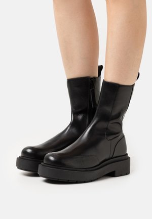 Bottes noires en cuir à la cheville avec une texture lisse, bout rond, semelle épaisse et zip latéral pour un enfilage facile.