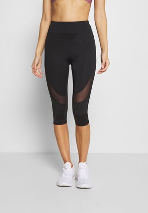 Frau trägt schwarze kurze Leggings mit durchsichtigen Netzpaneelen und weiße Sportschuhe, steht vor einem einfarbigen Hintergrund.