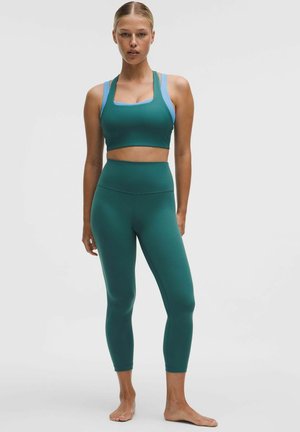 Femeie stând desculță, purtând colanți de culoare teal cu talie înaltă și un top scurt asortat, cu abdomenul expus, pe un fundal simplu.