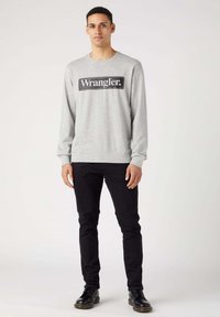 Šedá mikina s dlouhými rukávy, na které je černý obdélníkový logo "Wrangler". Vybaveno úzkými černými džíny a černými šněrovacími botami.