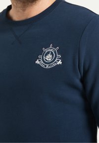 Granatowa bluza wykonana z miękkiego materiału, z okrągłym dekoltem i haftowanym na piersi białym i jasnoniebieskim logo w kształcie koła sterowego.