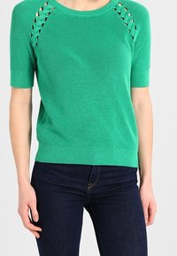 Grøn kortærmet strikket sweater med udskæringer på skuldrene, ribbet tekstur og tætsiddende nederkant. Sammen med mørke denim jeans.