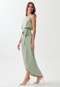 Dolga, svetlo zelena satenska obleka z oprijetim vrhom, detajlom na pasu v obliki mašne in s sproščenim krilom. Ima visok razporek in tanke trakaste petke.