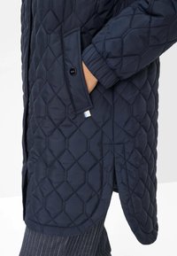 Marineblå quiltet lang frakke med diamantmønster, sidelommer og ribbede manchetter. Har et lille farvet mærke nær kanten.
