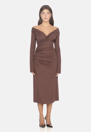 Vestito marrone off-shoulder con maniche lunghe, dettaglio arricciato sul davanti e una silhouette aderente, realizzato in un tessuto liscio. Indossato con tacchi alti scuri.