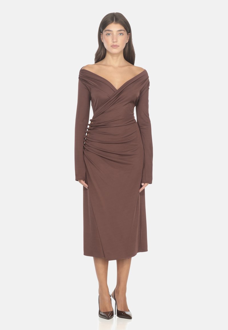 Vestito marrone off-shoulder con maniche lunghe, dettaglio arricciato sul davanti e una silhouette aderente, realizzato in un tessuto liscio. Indossato con tacchi alti scuri.