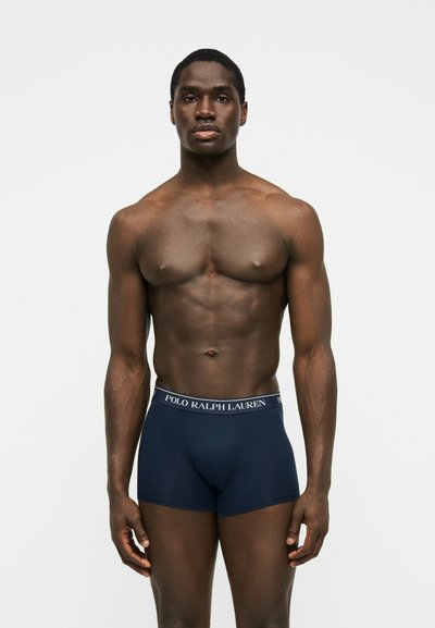 Homme musclé à la peau foncée, debout face à l’avant, portant un boxer Polo Ralph Lauren bleu marine sur un fond blanc uni.