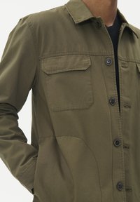 Olivno zelena srajca s poklopcem, dva žepa na prsih in gladko, teksturirano blago. Ima črne gumbe in krojen fit.