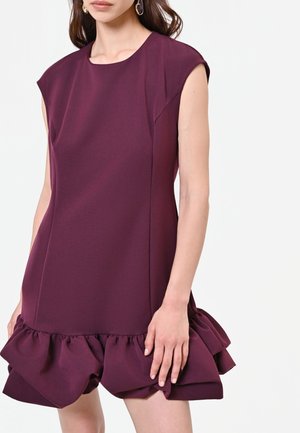 Femme portant une robe sans manches violet foncé avec un col rond et un ourlet à volants, debout devant un fond blanc uni.