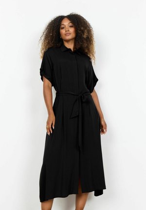 Kvinde med krøllet hår iført en sort midi-kjole med korte ærmer, krave og bindebælte i taljen, stående foran en hvid baggrund.