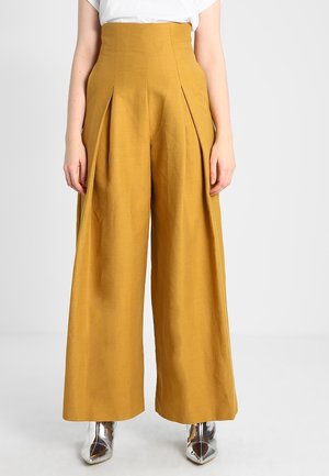 Vrouw draagt een mosterdgele broek met hoge taille en brede pijpen met plooien, gecombineerd met een wit topje en zilveren puntige laarzen.