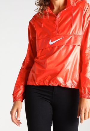 Femme portant une veste coupe-vent Nike rouge vif avec une fermeture éclair frontale et des leggings noirs sur un fond blanc uni.