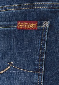 Jeans de mezclilla azul oscuro con una etiqueta roja y texto amarillo. Costuras doradas notables a lo largo de las costuras y un botón metálico como acento.