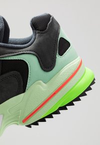 Kengät, joissa on musta verkkopinta, mintunvihreät mokkanahkaosuudet, kirkas punainen raita välipohjassa, teksturoitu vihreä ulkopohja kolmioharjanteilla.