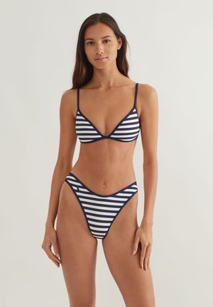 Ženska z dolgimi temnimi lasmi, oblečena v mornarsko modro-beli bikini z ozkimi naramnicami, stoji pred enostavnim svetlim ozadjem.