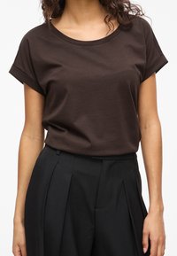 Kvinde iført en mørkebrun kortærmet t-shirt stoppet ned i sorte plisserede bukser med høj talje, vist fra hals til hofter.