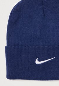 Tengerészkék kötött sapka hajtott gallérral és fehér hímzett Nike logóval. Az anyag texturált megjelenésű és szorosan illeszkedik.