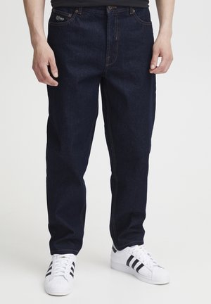 Jeans in denim blu scuro con taglio dritto, con cuciture arancioni. Abbinati a sneakers bianche con strisce nere.