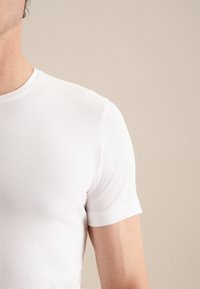 T-shirt blanche ajustée à manches courtes, en coton lisse, avec un col rond et des coutures minimales, mettant en avant un design épuré et simple.