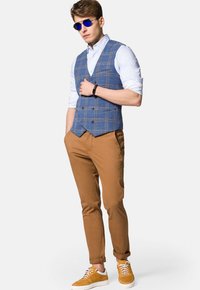 Niebieska kraciasta kamizelka na białej koszuli, brązowe chinosy i żółte trampki. Dodatki to okrągłe niebieskie okulary przeciwsłoneczne oraz smukły czarny zegarek.