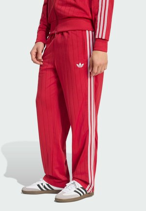 Osoba ubrana w czerwone spodnie dresowe i kurtkę Adidas z białymi paskami, stojąca z rękami swobodnie opuszczonymi wzdłuż ciała.
