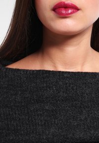Maglione nero lavorato a maglia con scollo ampio, tessuto testurizzato e vestibilità morbida. Lo sfondo è neutro, evidenziando i dettagli del capo.