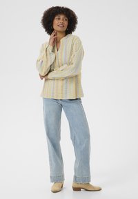 Chemise à manches longues rayée de couleur jaune clair, avec des boutons sur le devant, présentant des motifs texturés. Portée avec un jean large bleu clair et des bottes beiges.