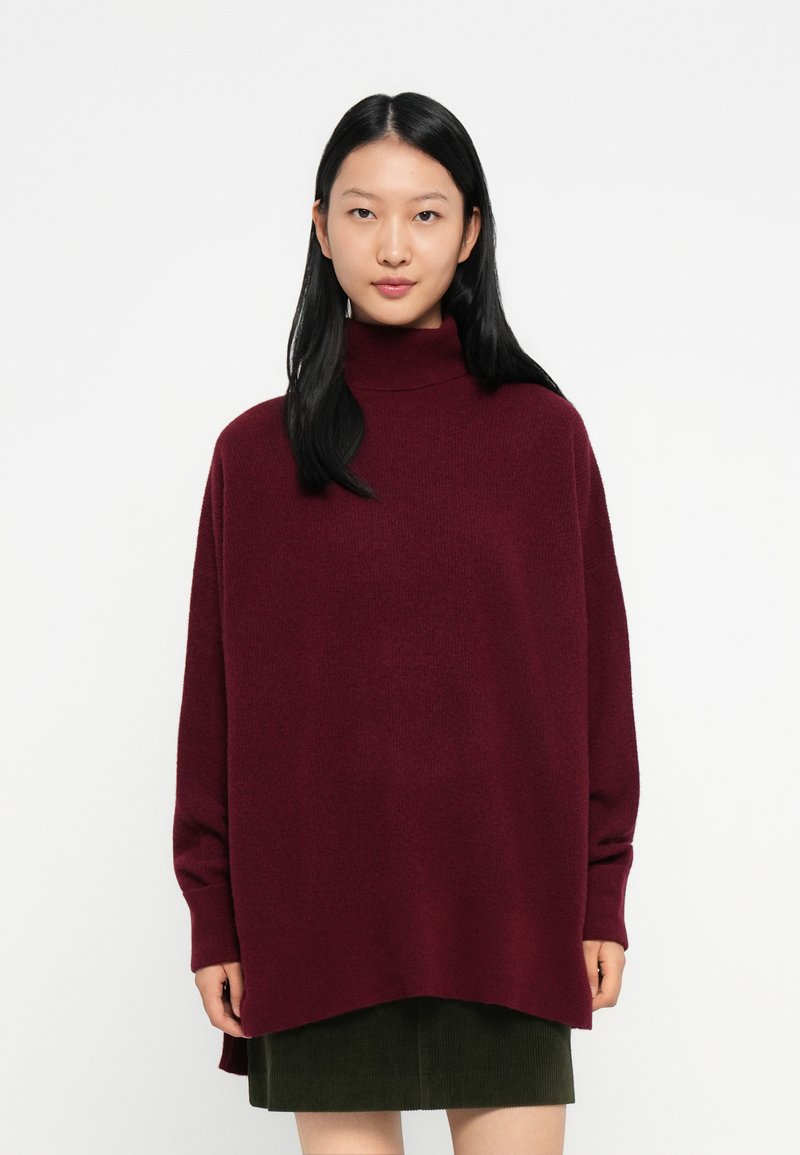 Mlada ženska z dolgimi črnimi lasmi, oblečena v ohlapen burgundski pulover s turtleneckom in temno zeleno krilo, ki stoji ob belem ozadju.