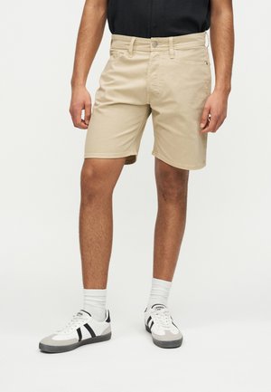 Mężczyzna w beżowych szortach do kolan, białych trampkach z czarnymi paskami, białych skarpetkach i czarnej koszulce na jednolitym tle.