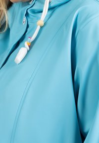 Světlomodrý pláštěnka s lesklým povrchem, vybavená zipovým uzávěrem, kapucí se stahovací šňůrkou a bílým třásňovým detailem.