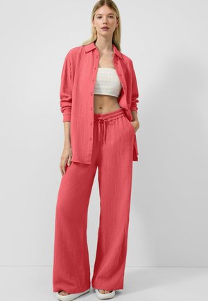 Kobieta ubrana w koralowe, teksturowane spodnie z szerokimi nogawkami, dopasowaną koszulę na guziki, biały szydełkowany krótki top i białe sandały, stoi na szarym tle.