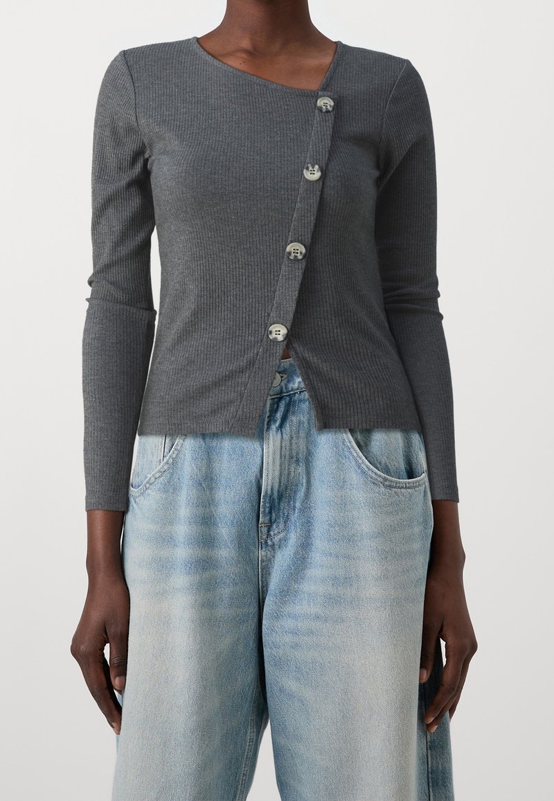 Femme portant un haut gris ajusté côtelé à manches longues avec une fermeture asymétrique à boutons, associé à un jean ample délavé clair.
