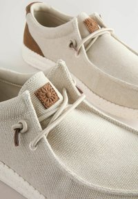 Beige canvas kengät ruskeilla nahkaakkosilla, pyöreät nauhat, teksturoitu päällinen ja valkoispilkkuinen pohja. Sivulla on neliönmuotoinen logopatch.
