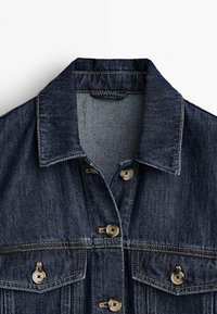 Veste en denim bleu foncé avec col, fermeture à boutons sur le devant et deux poches poitrine boutonnées, présentée sur fond blanc.