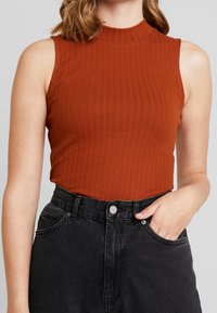 Kvinna i ärmlös ribbad topp i rostfärg som är stoppad i högmidjade svarta jeans med handen i fickan på enfärgad bakgrund.