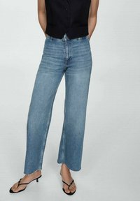 Vaqueros de mezclilla azul claro de pierna ancha con dobladillo sin rematar, cintura alta y sutiles pliegues, combinados con un chaleco ajustado negro y sandalias de tiras.