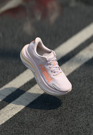 Chaussure de course Hoka blanche et orange en l'air au-dessus d'une route en asphalte avec des marquages de voie blancs, projetant une ombre en dessous.